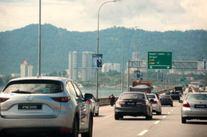 Busy day on Penang Bridge with cars traveling under a cloudy sky, featuring mountains and cityscape.